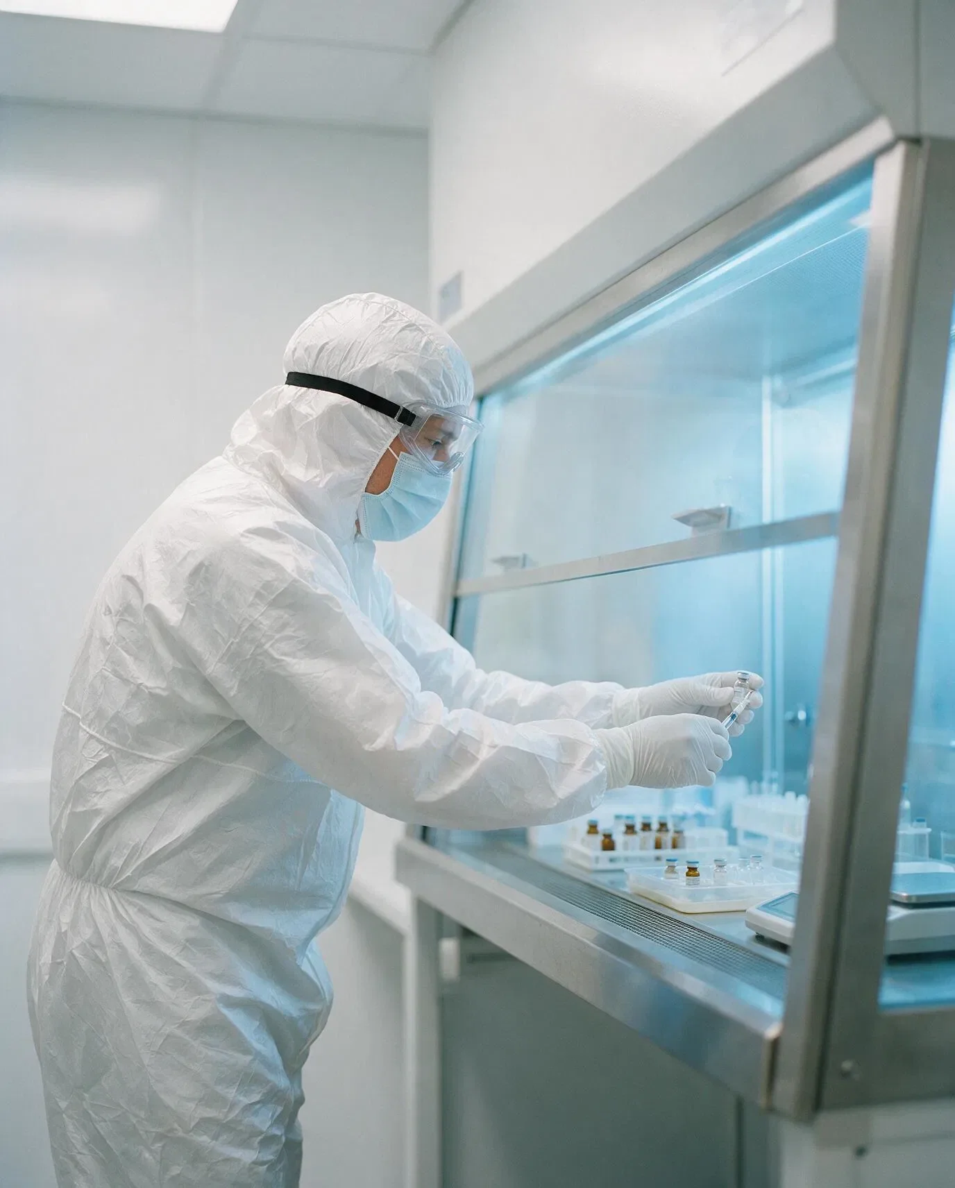 A technician in a sterile gown, goggles and gloves handles a small vial inside a laminar airflow hood